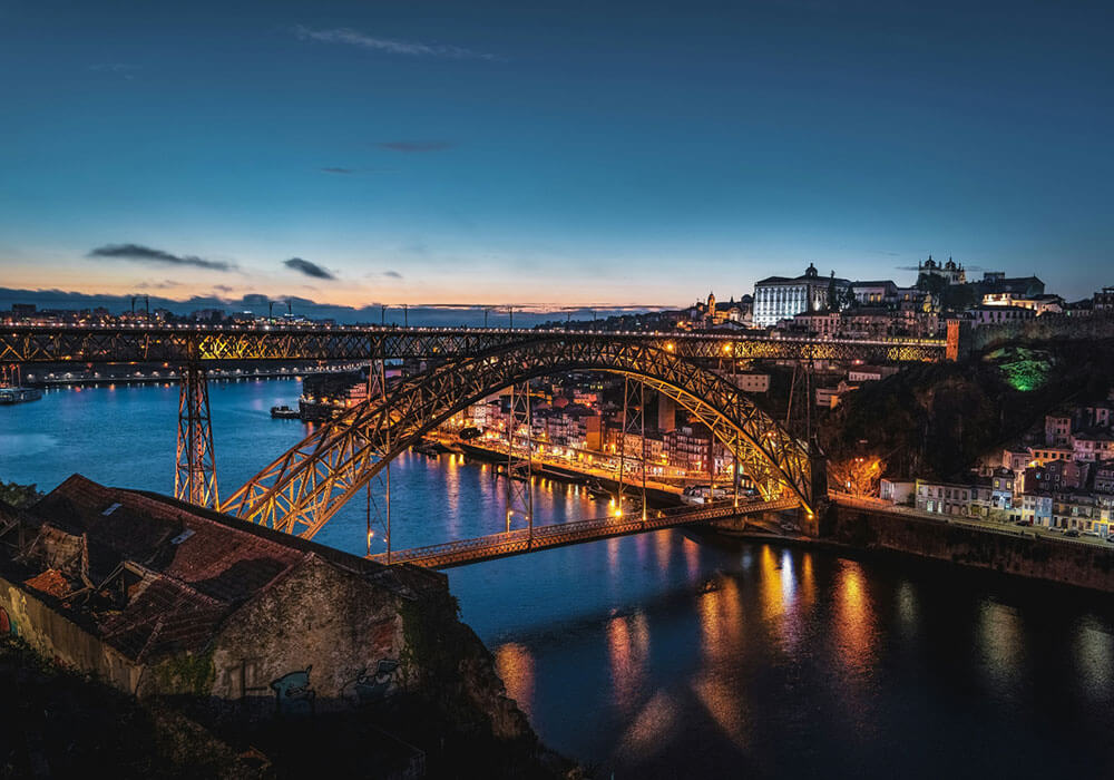 Panoramic view of Luís I Bridge at sunset, illuminated over the Douro River, with the city of Porto in the background.